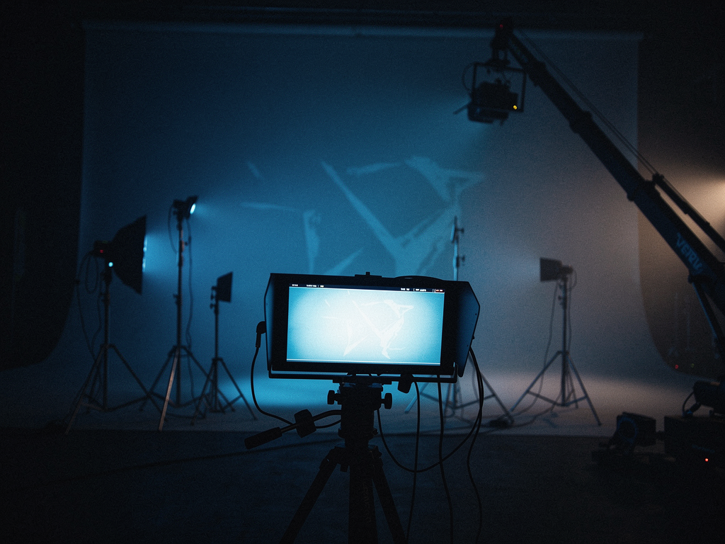 A film set in low-key blue light: camera monitor in the foreground, crane and lighting rigs in the haze