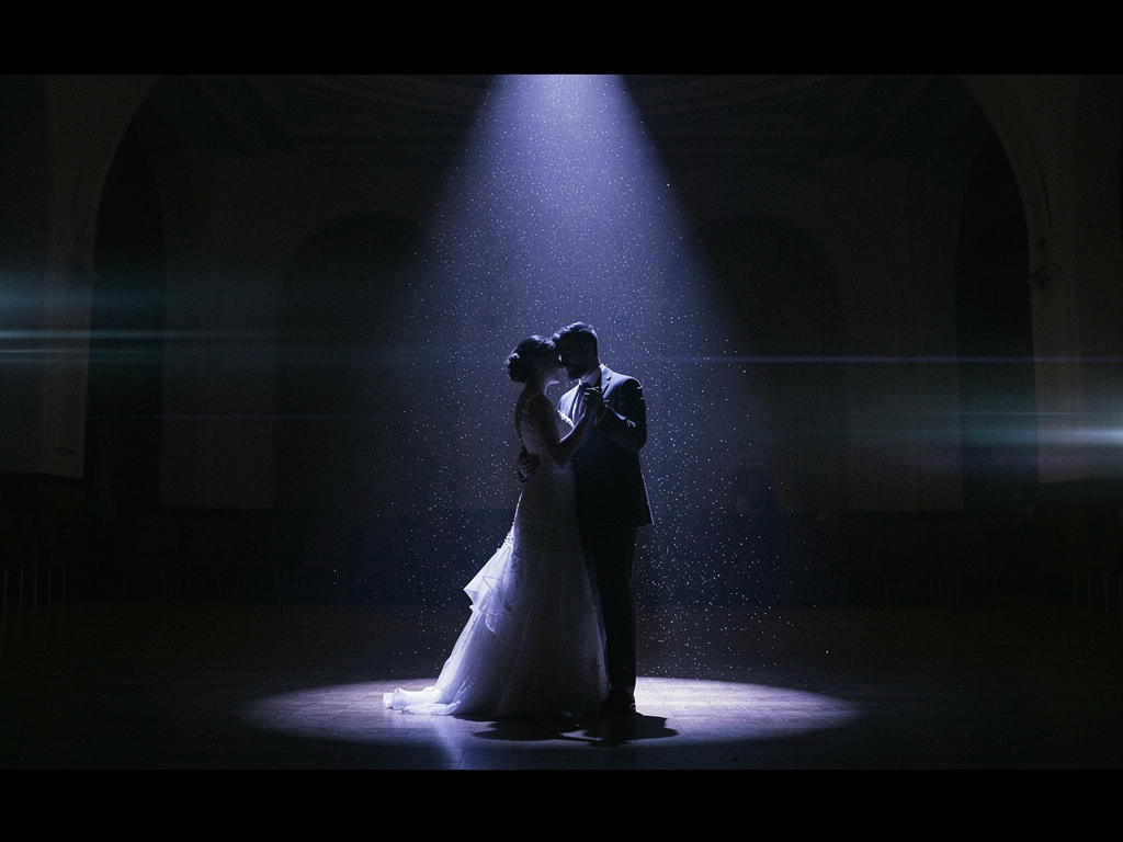 A bride and groom dancing alone under a single theatrical spotlight in a dark hall